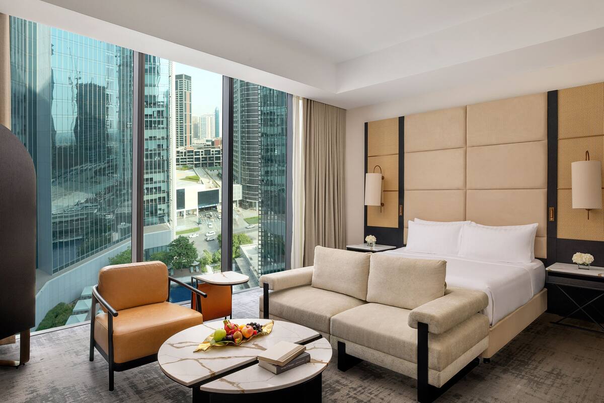 Habitación de hotel moderna con una cama grande, un sofá, una mesita de café y vistas de la ciudad a través de ventanales de piso a techo.