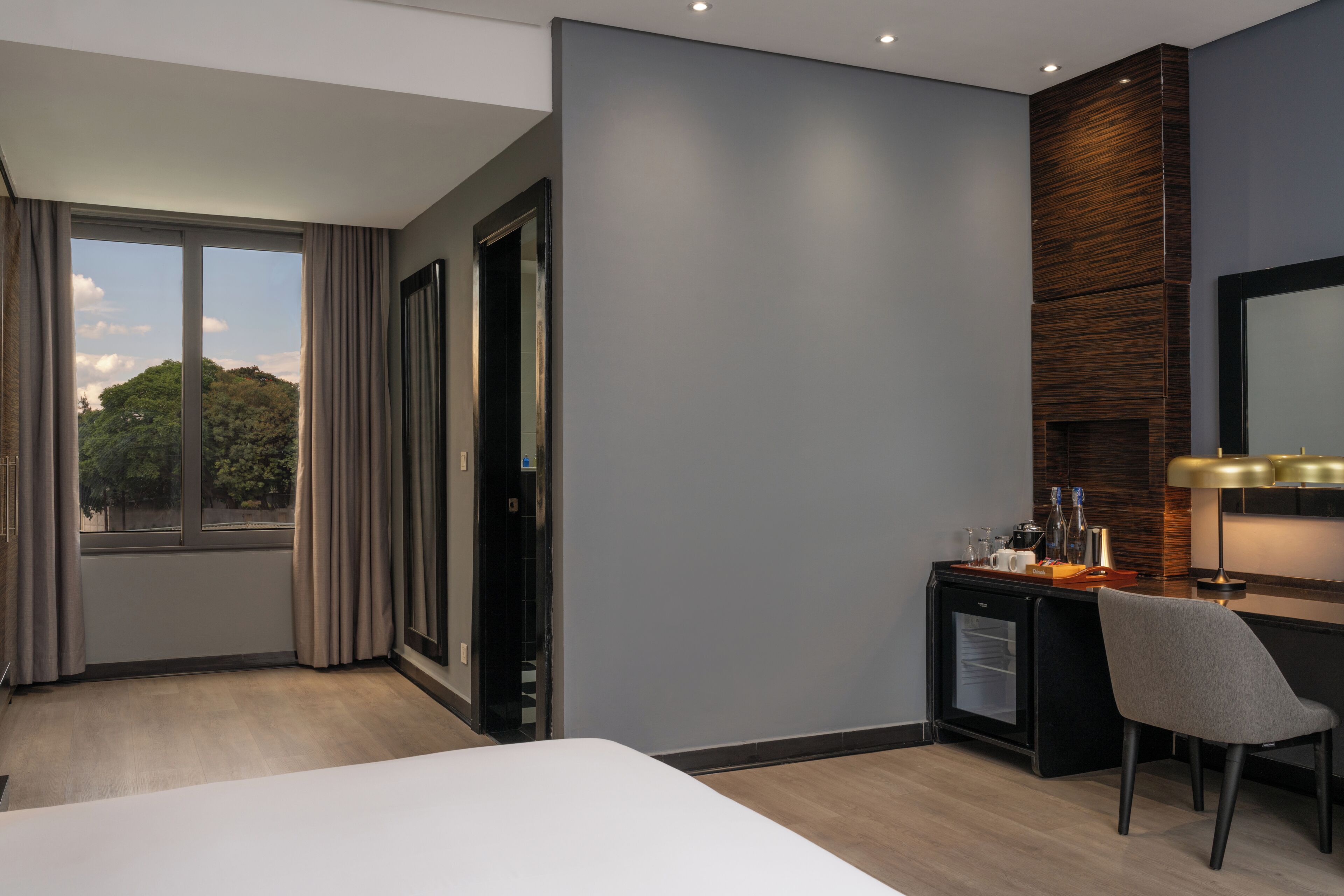A modern hotel room with a bed, a desk with a chair, a minibar, and a large window with curtains.