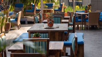 Een terras met houten tafels en stoelen, blauwe kussens en potplanten.