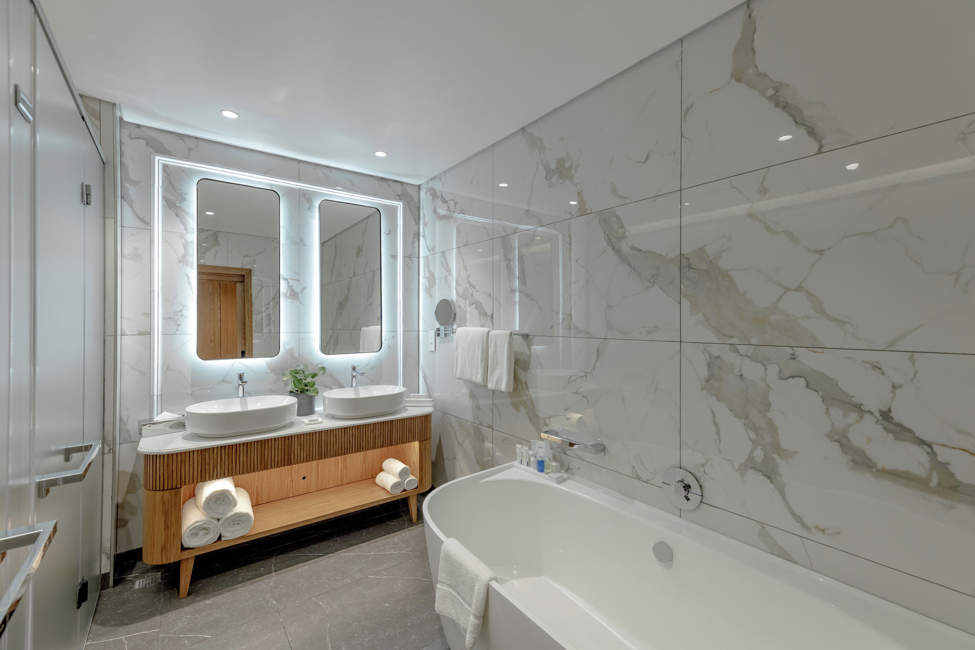 A modern bathroom with a large mirror, two sinks, a wooden vanity, a bathtub, and marble walls.