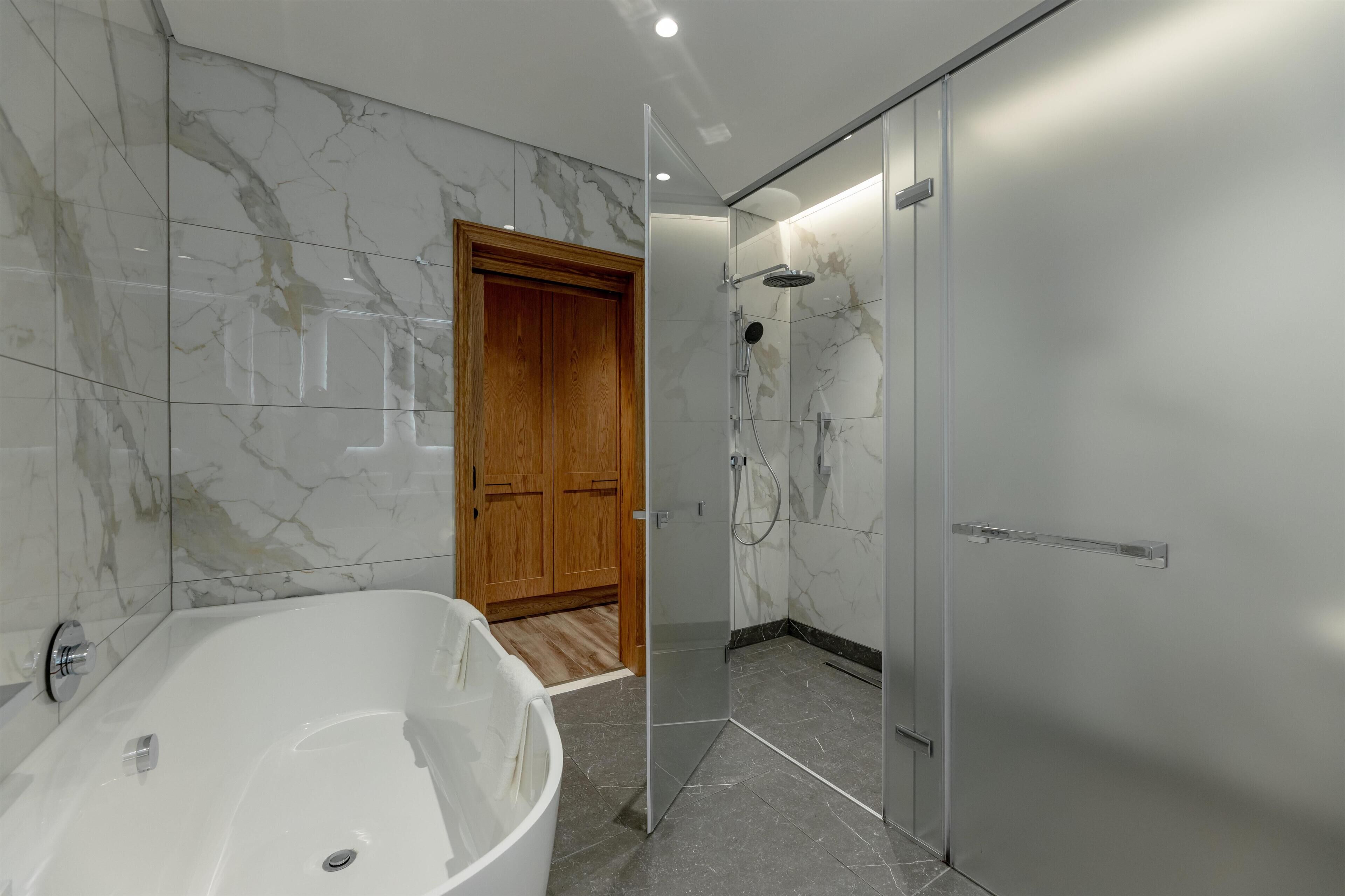 A modern bathroom with a marble floor, a freestanding bathtub, a glass-enclosed shower, and a wooden door.