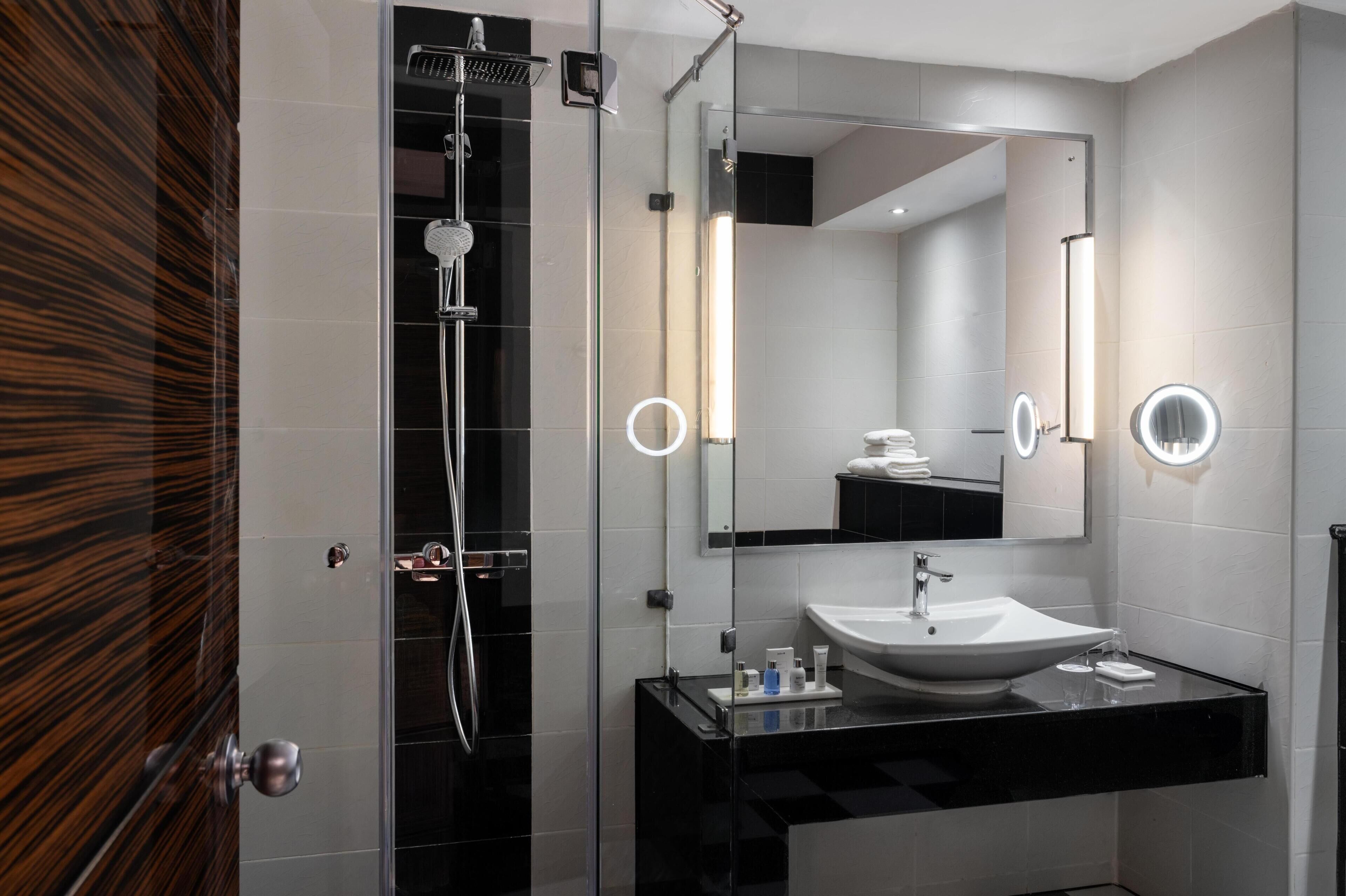 A modern bathroom with a glass-enclosed shower, a large mirror with built-in lights, and a black countertop with a white sink.