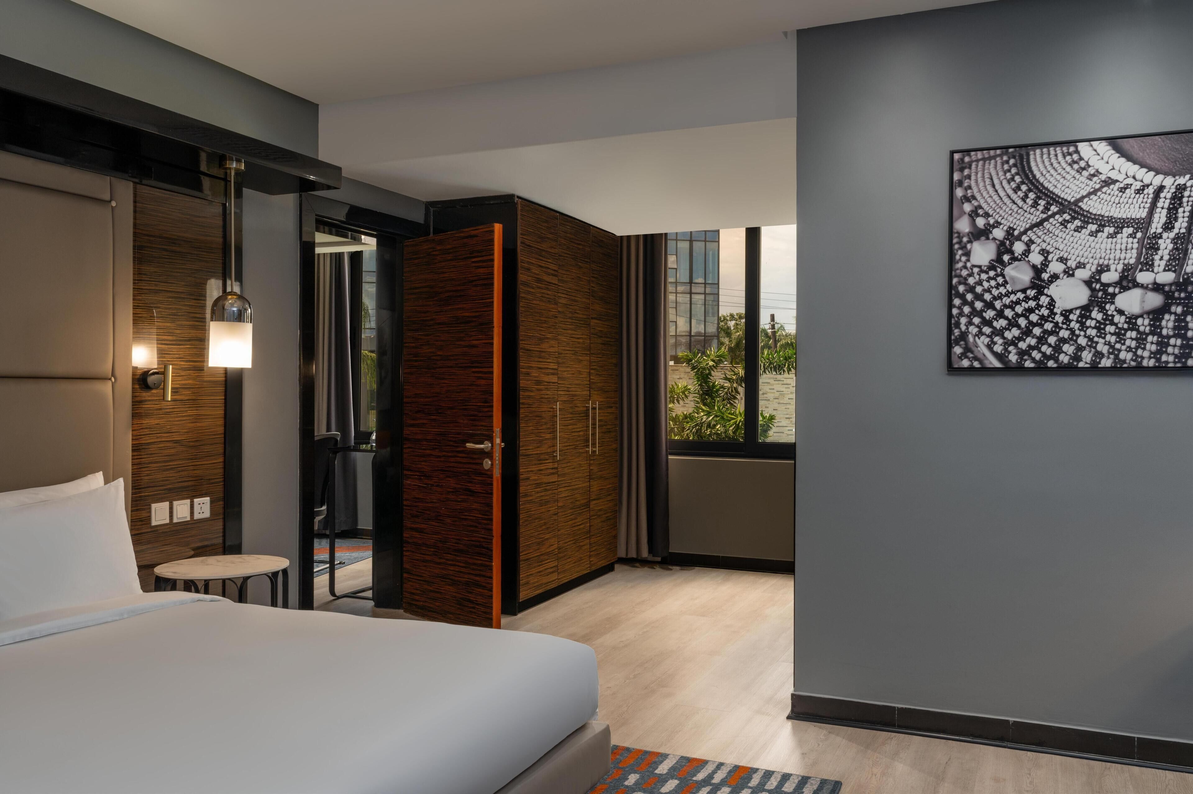 A modern hotel room with a large bed, a wooden wardrobe, a small table, and a large window with a view of greenery.
