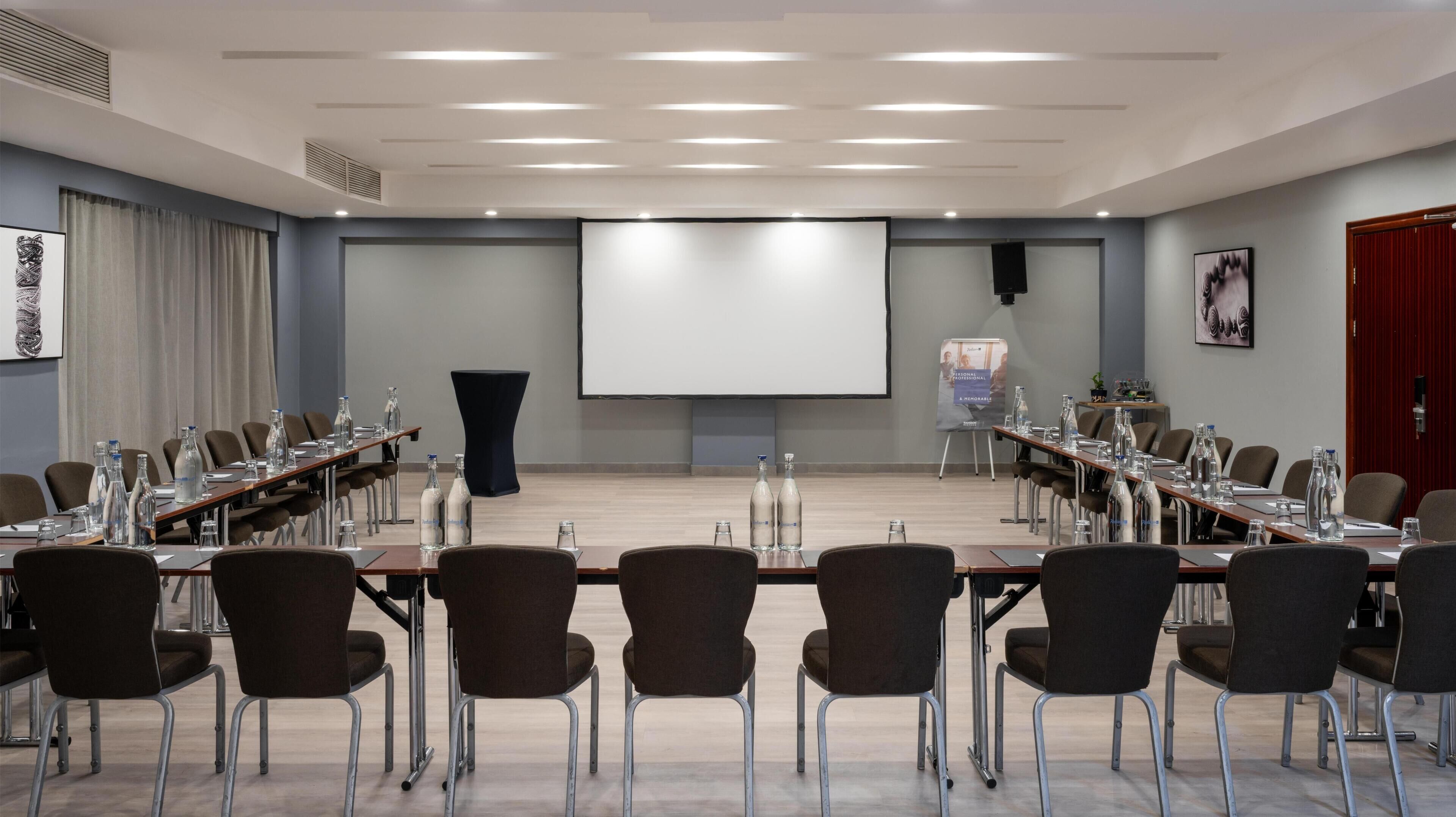 A conference room with a long table, chairs, water bottles, and a projector screen.