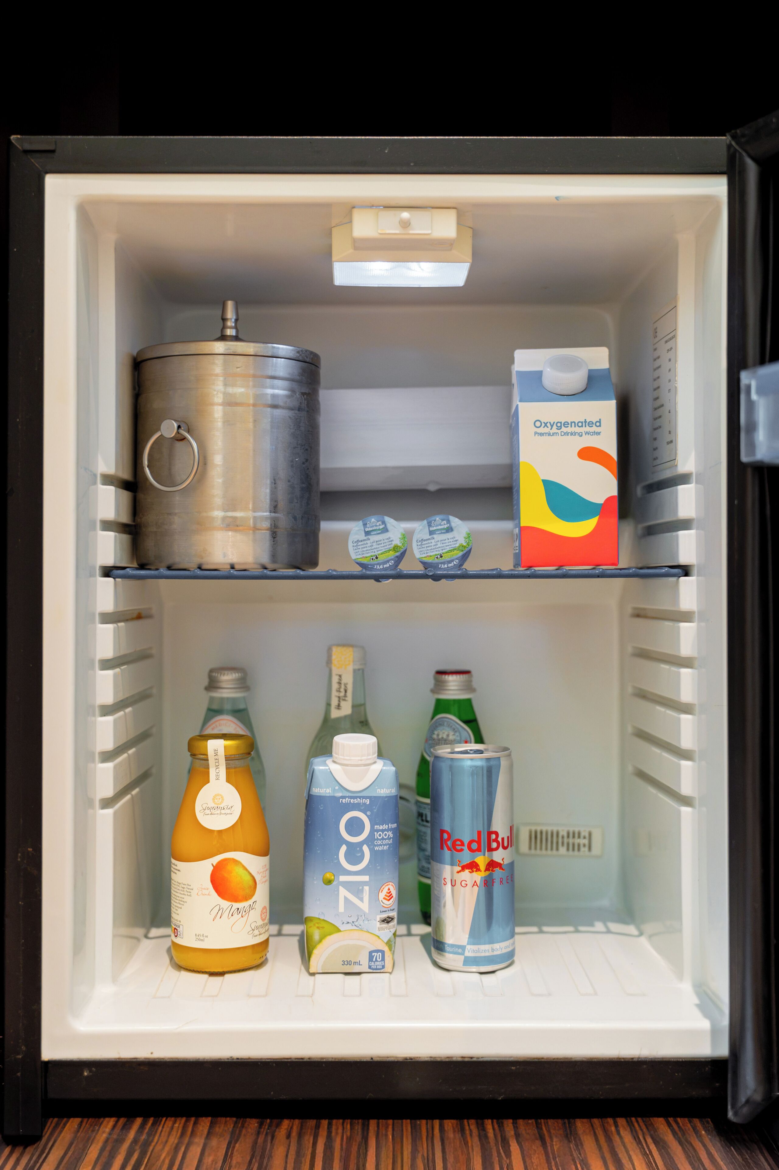 A refrigerator containing various beverages including a carton of Oxygenated Premium Drinking Water, a bottle of Mango juice, a carton of ZICO, and a can of Red Bull.