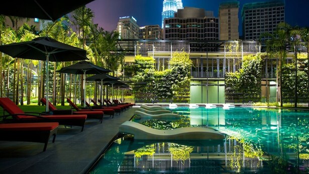 A pool area with lounge chairs and umbrellas, surrounded by greenery and buildings with illuminated facades. - E&O Residences Kuala Lumpur (Kuala Lumpur)