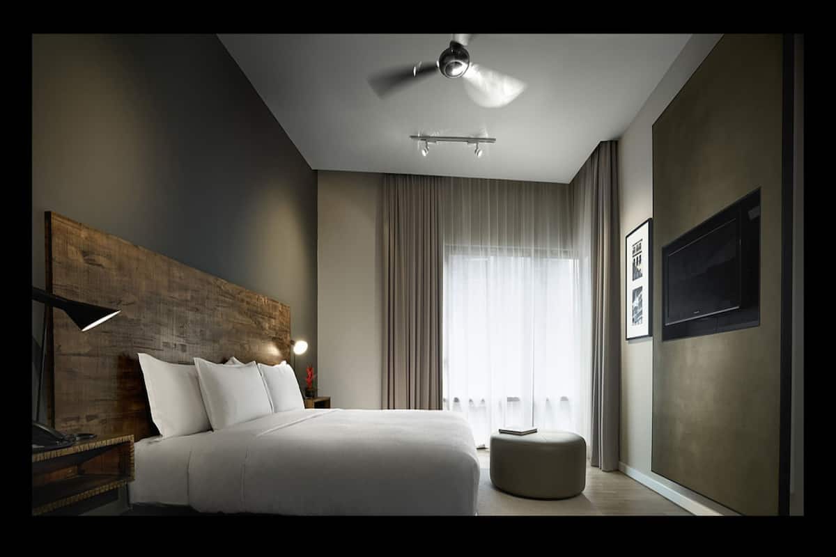 A modern hotel room with a large bed, a ceiling fan, a nightstand, a reading lamp, a window with curtains, and a framed picture on the wall.