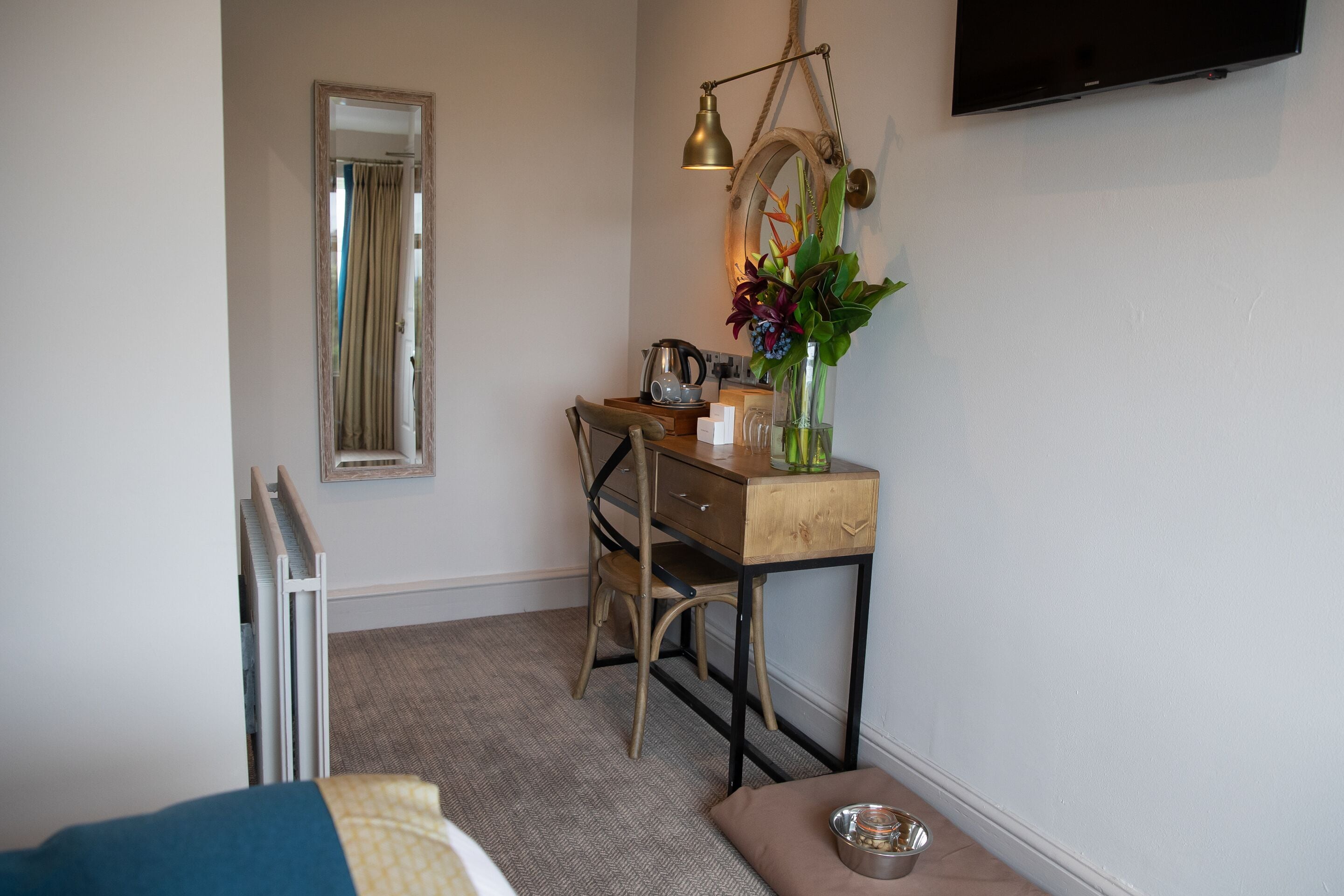 A compact room with a bed, a desk with a chair, a mirror, a TV, and a small table with a vase of flowers.