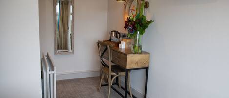 A compact room with a bed, a desk with a chair, a mirror, a TV, and a small table with a vase of flowers.