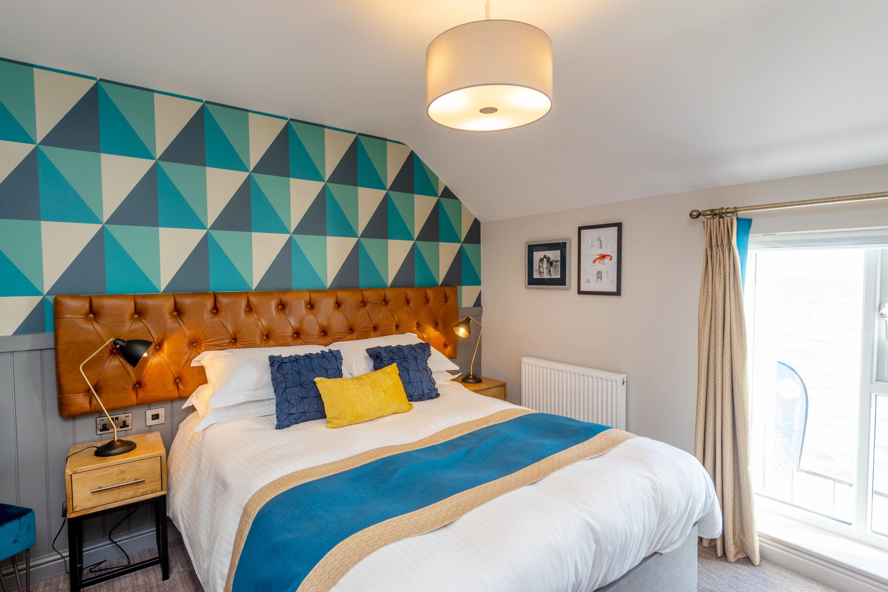 Un dormitorio moderno con un cabecero de estampado geométrico, una cama grande con colcha azul y beige, una mesita de noche con lámpara y una ventana con cortinas.