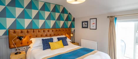 A modern bedroom with a geometric-patterned headboard, a large bed with a blue and beige bedspread, a nightstand with a lamp, and a window with curtains.