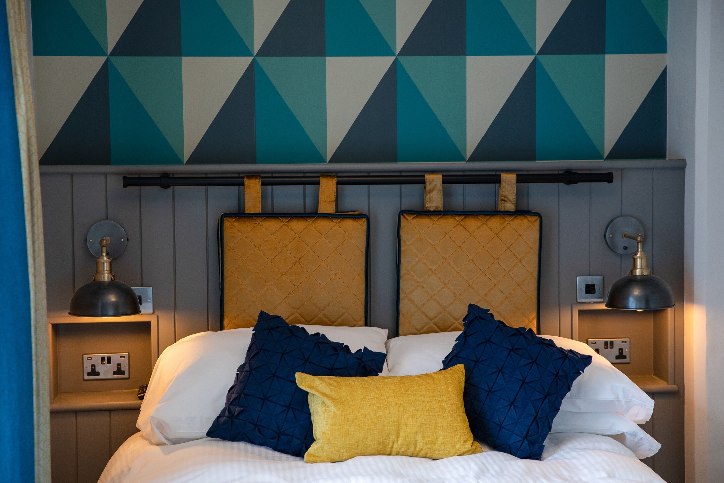 A neatly made bed with a headboard, two bedside tables, and a geometric-patterned wall above.