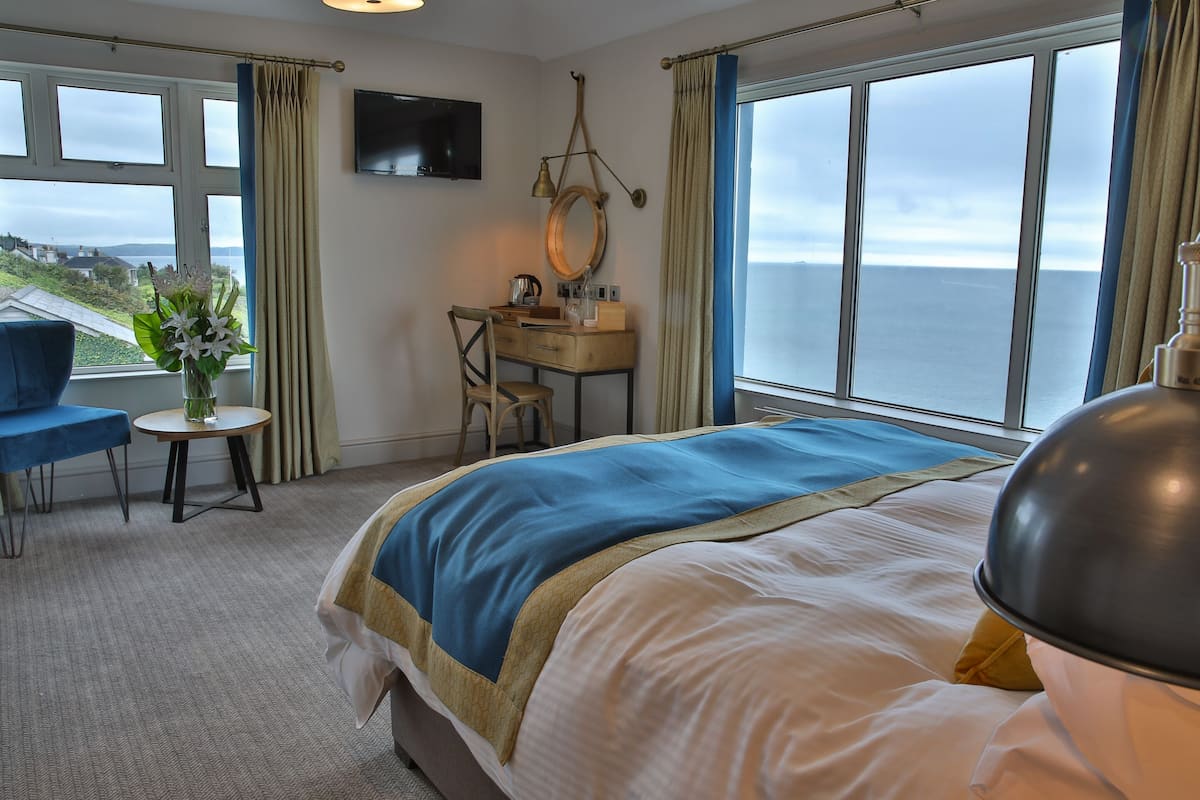 A hotel room with a bed, a desk, a chair, a TV, and a view of the sea.