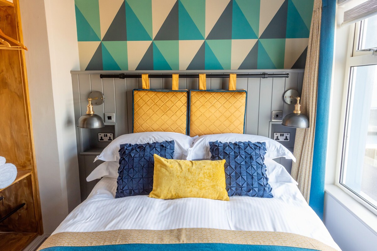 A neatly made bed with white and blue bedding, a yellow pillow, and a geometric-patterned headboard.