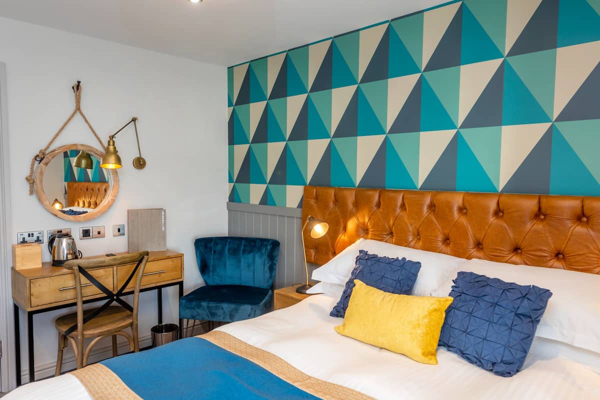 A hotel room with a tufted headboard, a bed with blue and yellow pillows, a desk with a chair, a mirror, and a geometric-patterned wall.
