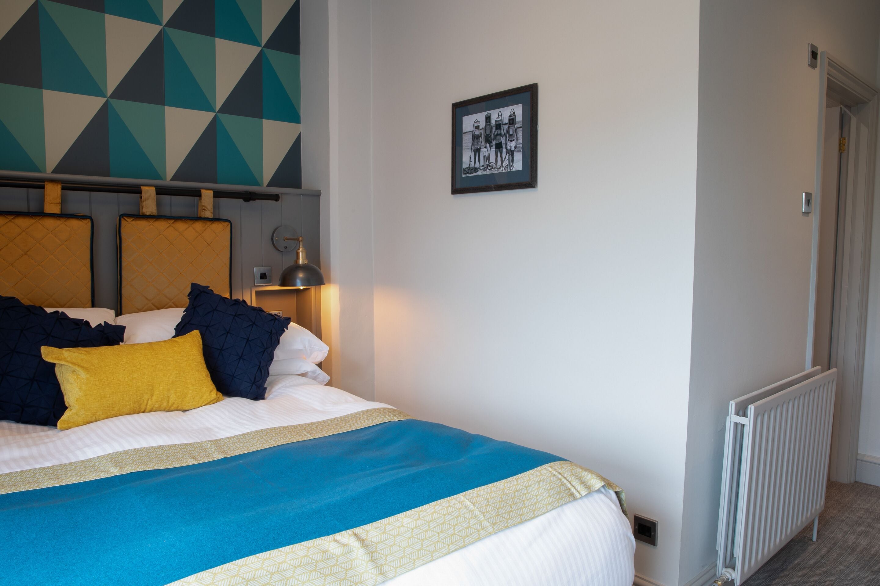 Una cama bien hecha con colcha azul y blanca, un almohadón amarillo y almohadas azul oscuro. Hay un cuadro en la pared y un radiador en el suelo.