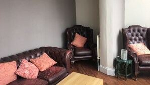 A living room with a dark brown leather sofa and two matching armchairs, each with a patterned cushion. A small green stool with a decorative item is placed beside one armchair. - The Kingscliff (Blackpool)