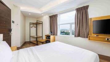 A hotel room with a bed, a TV, a wardrobe, a desk with a chair, a shelf, and a window with curtains.