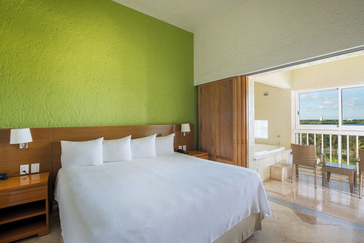 A hotel room with a large bed, wooden headboard, and a view of a balcony with a pool.