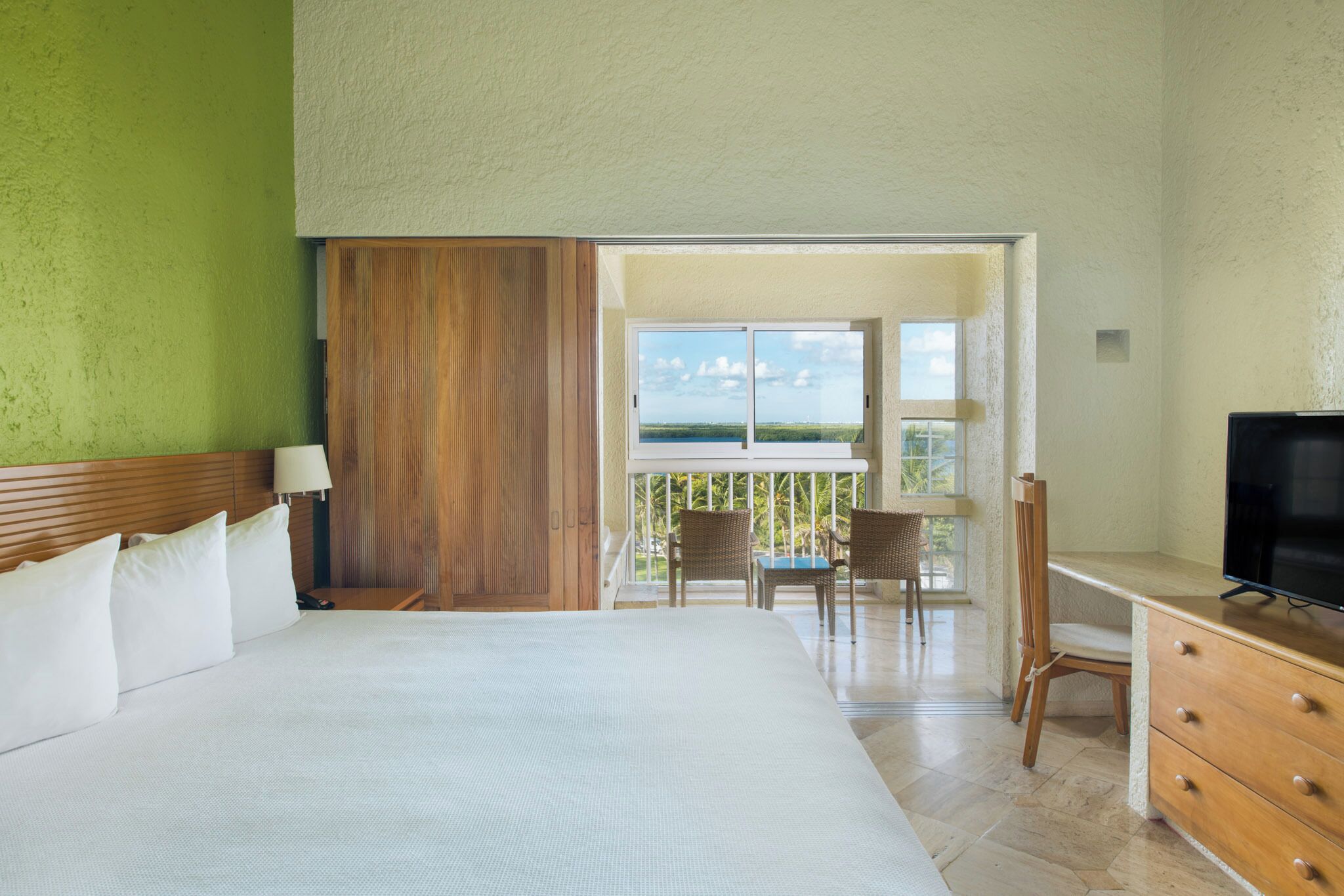 Habitación de hotel con cama, escritorio, televisor, balcón con sillas y vista al océano.