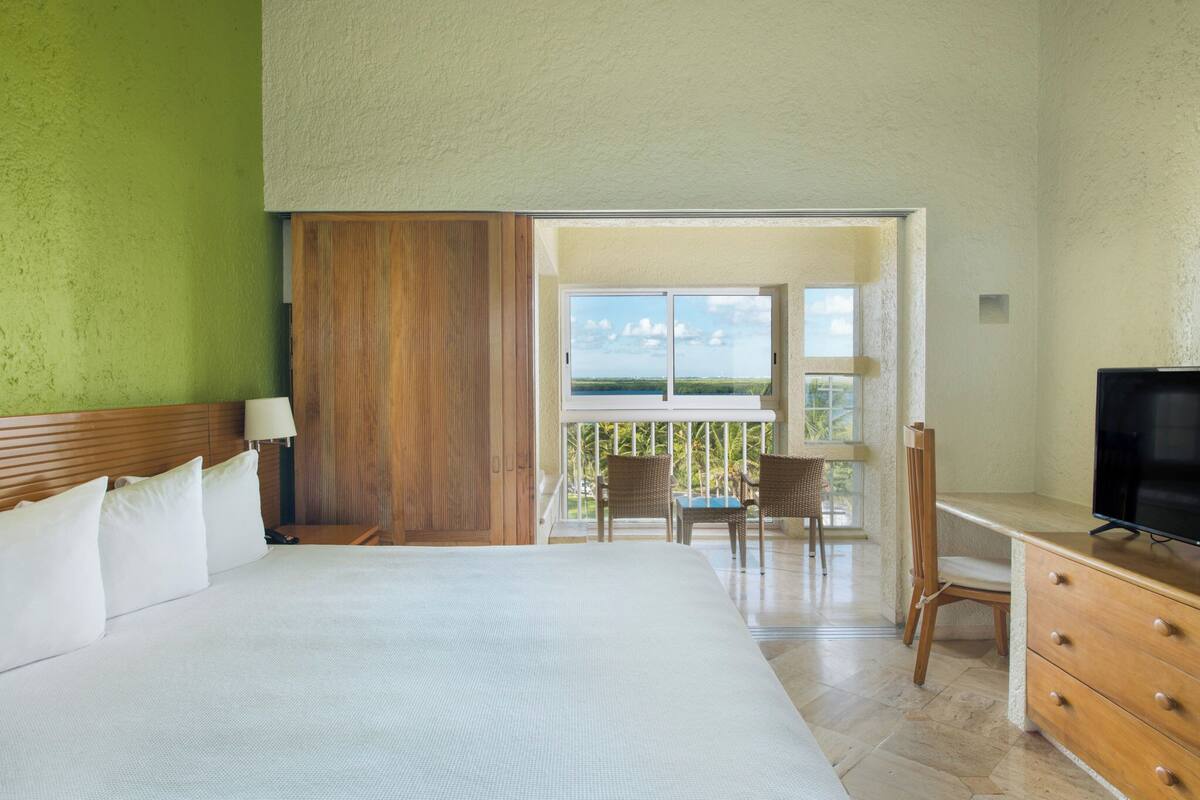 Habitación de hotel con cama, escritorio, televisor, balcón con sillas y vista al océano.