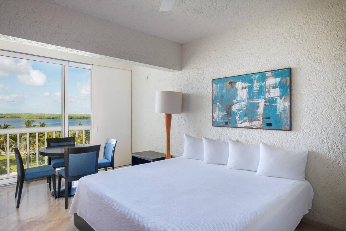 Habitación de hotel con cama, escritorio, silla, lámpara y una ventana grande con vista al océano.