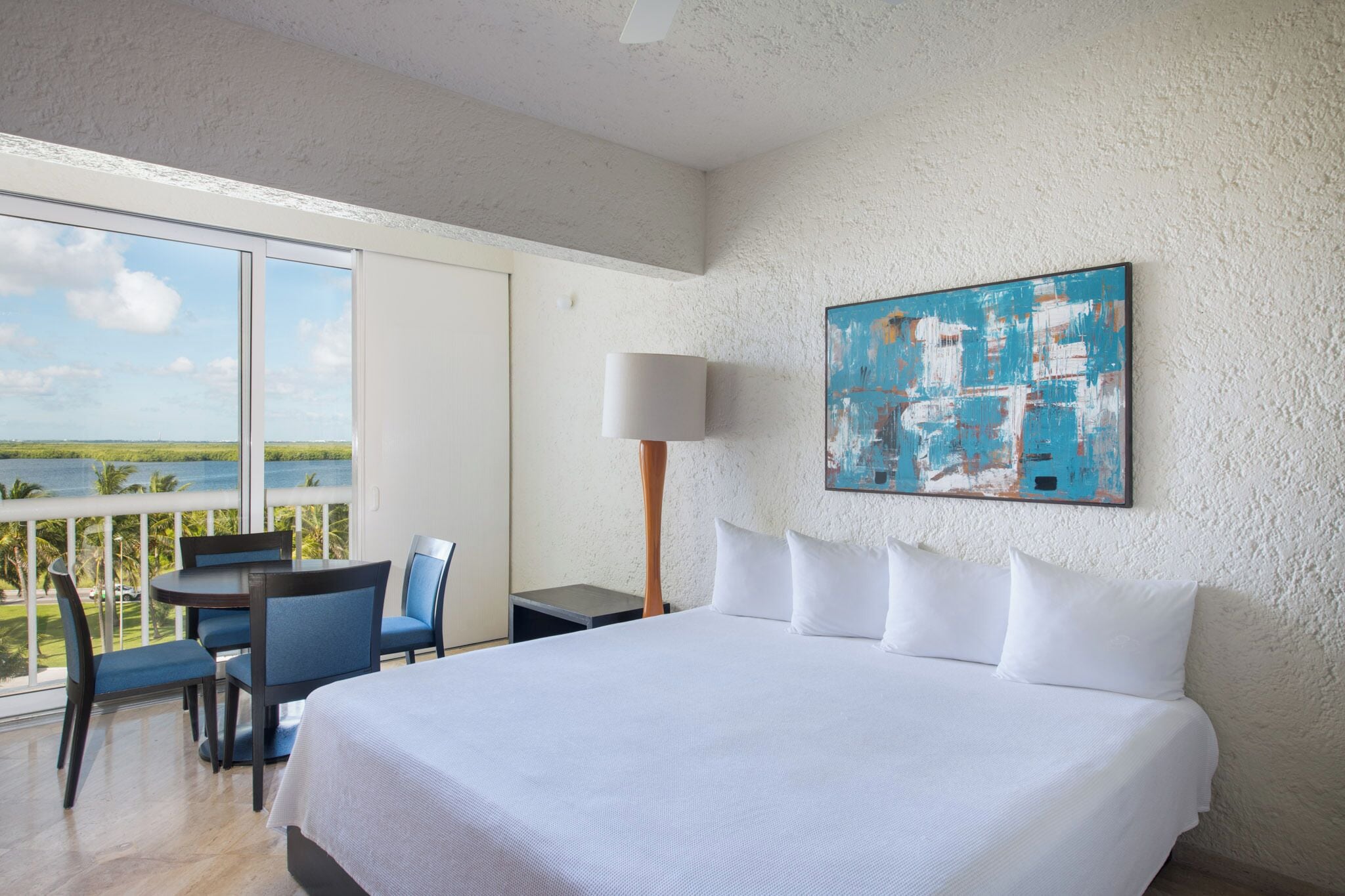 Habitación de hotel con una cama grande, un escritorio con silla, una lámpara y un cuadro en la pared.