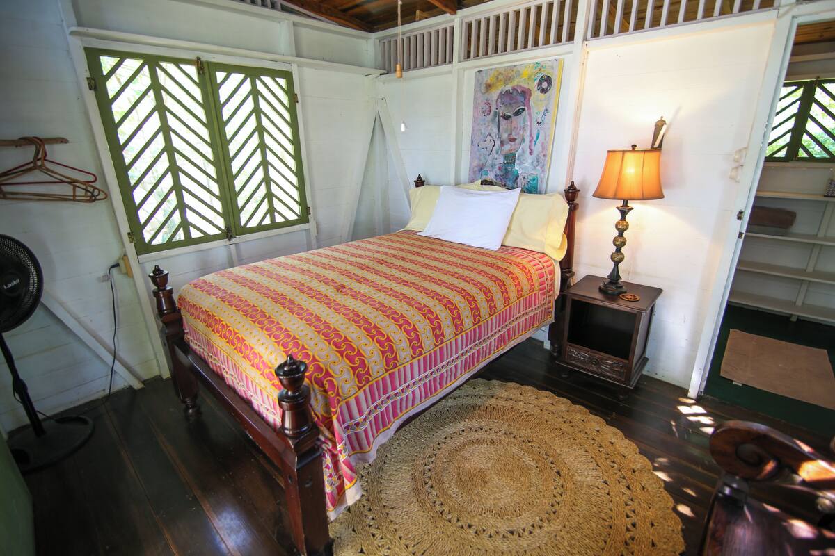 A room with a bed, a fan, a lamp, and a woven rug.