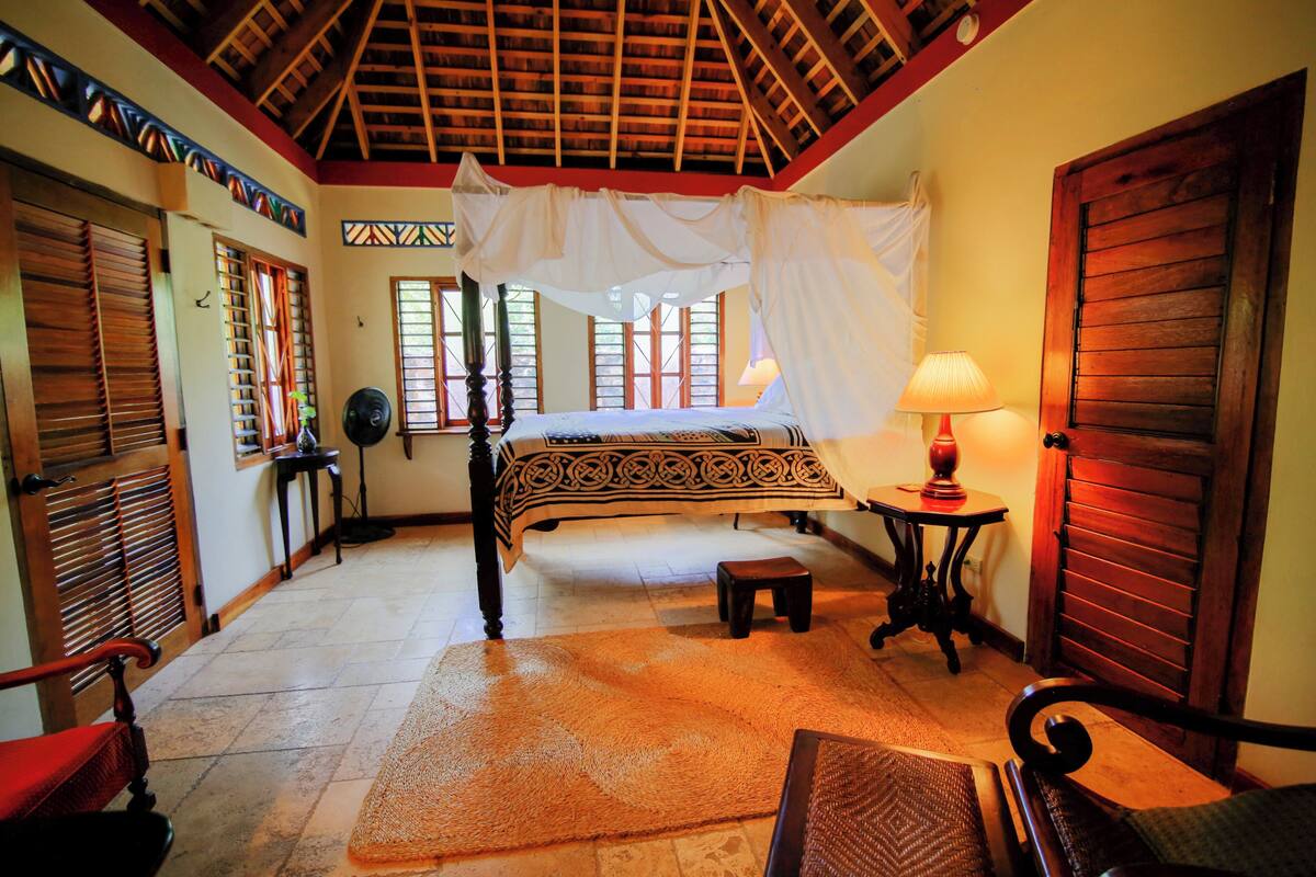A room with a bed, a chair, a table, and a fan.