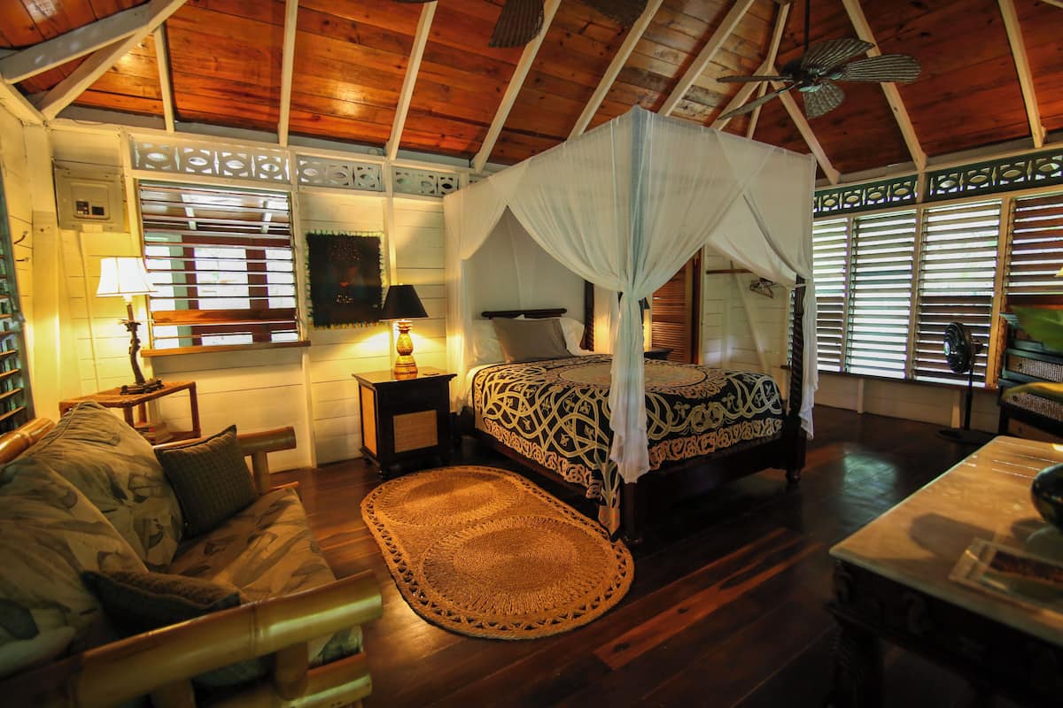 A room with a bed, a wooden ceiling, a ceiling fan, a nightstand, a lamp, a woven rug, and a window with blinds.