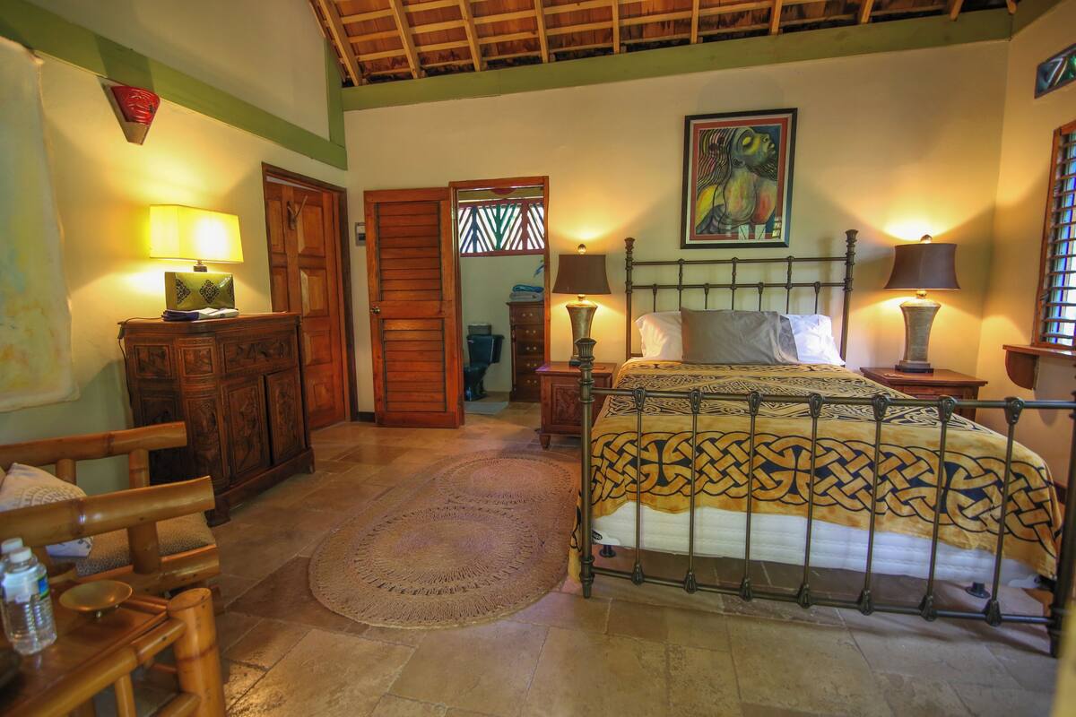 A bedroom with a bed, wooden furniture, a painting, and a door leading to another room.