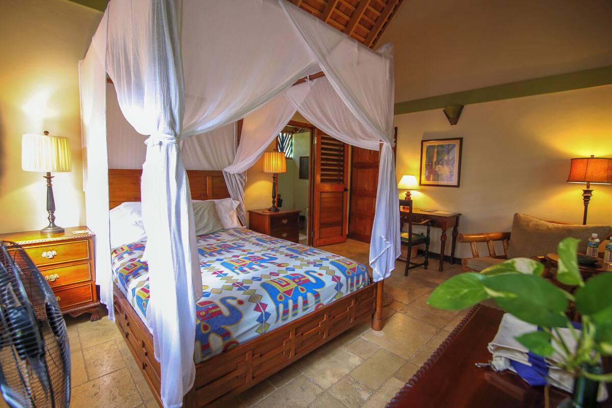 A four-poster bed with a colorful bedspread, a wooden nightstand with a lamp, a small table with a lamp, and a chair in a hotel room.