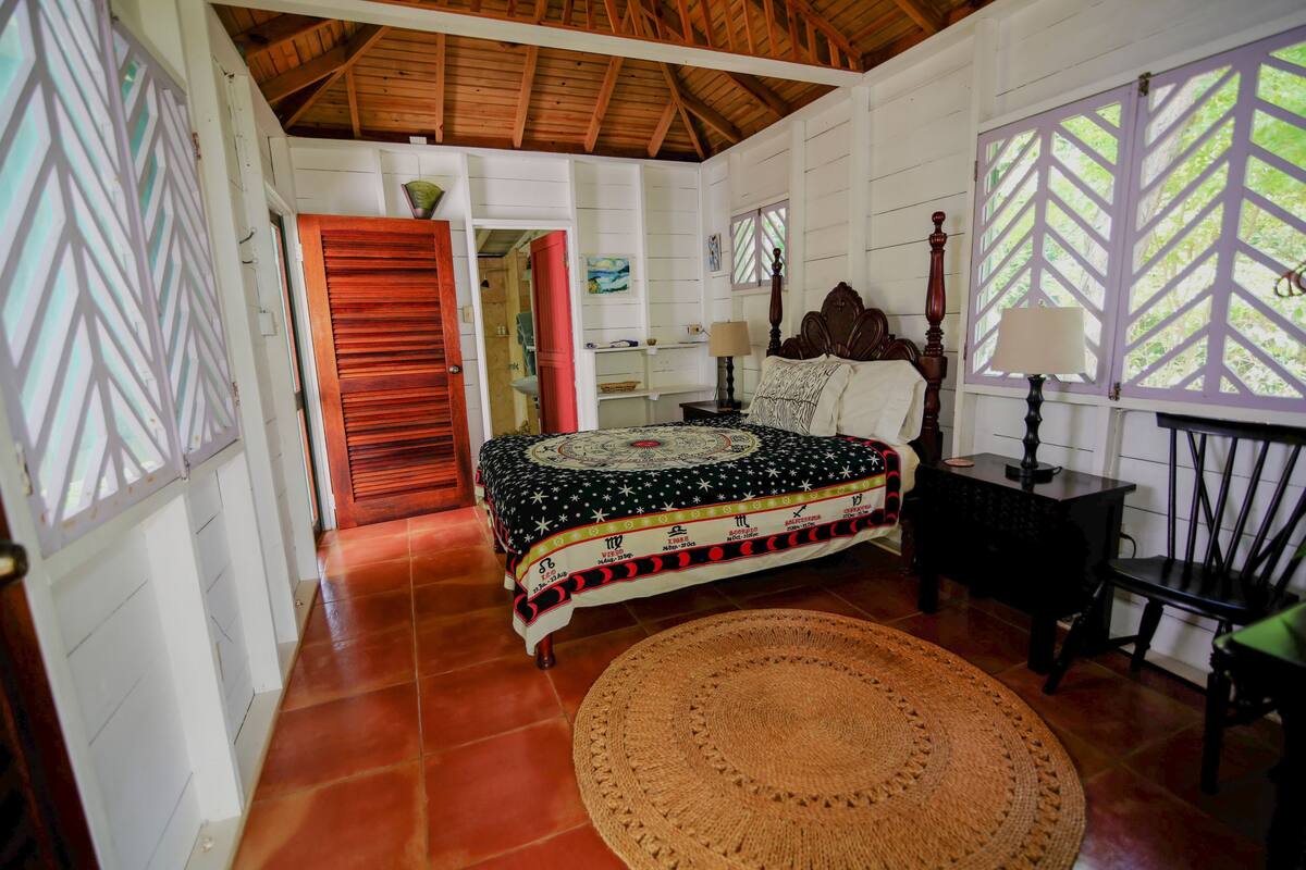 A room with a bed, a round woven rug, a nightstand, a chair, and a window with a lattice design.