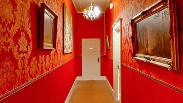 Un couloir aux murs rouges, orné d’un lustre et de tableaux encadrés.