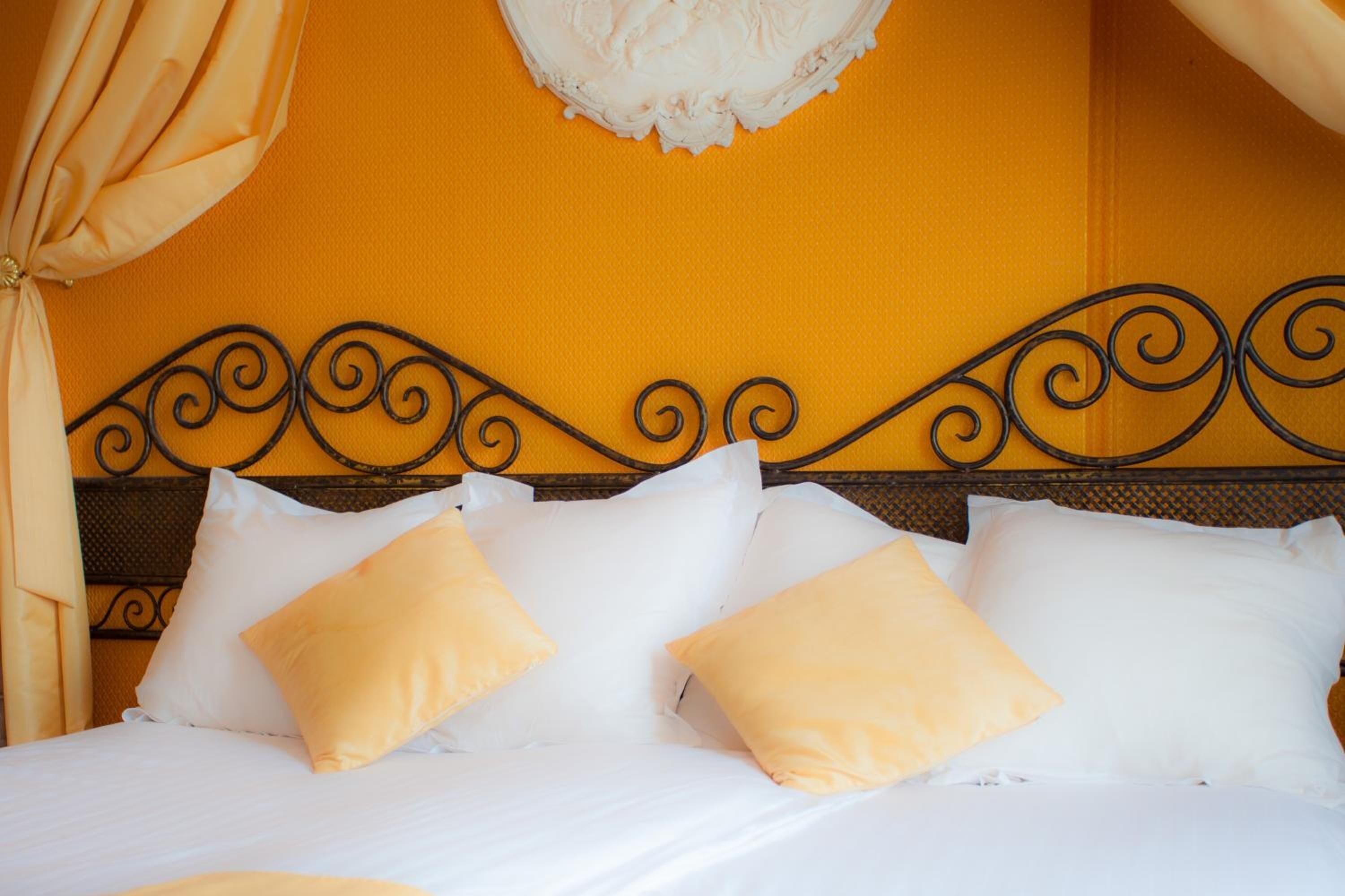 A bed with white linens and yellow pillows, a black metal headboard, and a yellow wall with a decorative white ceiling piece.