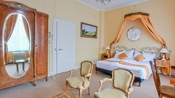 A hotel room with a large bed, a wardrobe, two armchairs, a small table, and a chandelier.