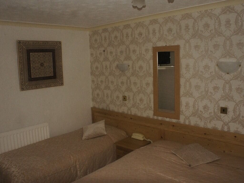 A room with two single beds, a wooden headboard, a patterned wallpaper, a framed picture, and a small bedside table with a telephone.