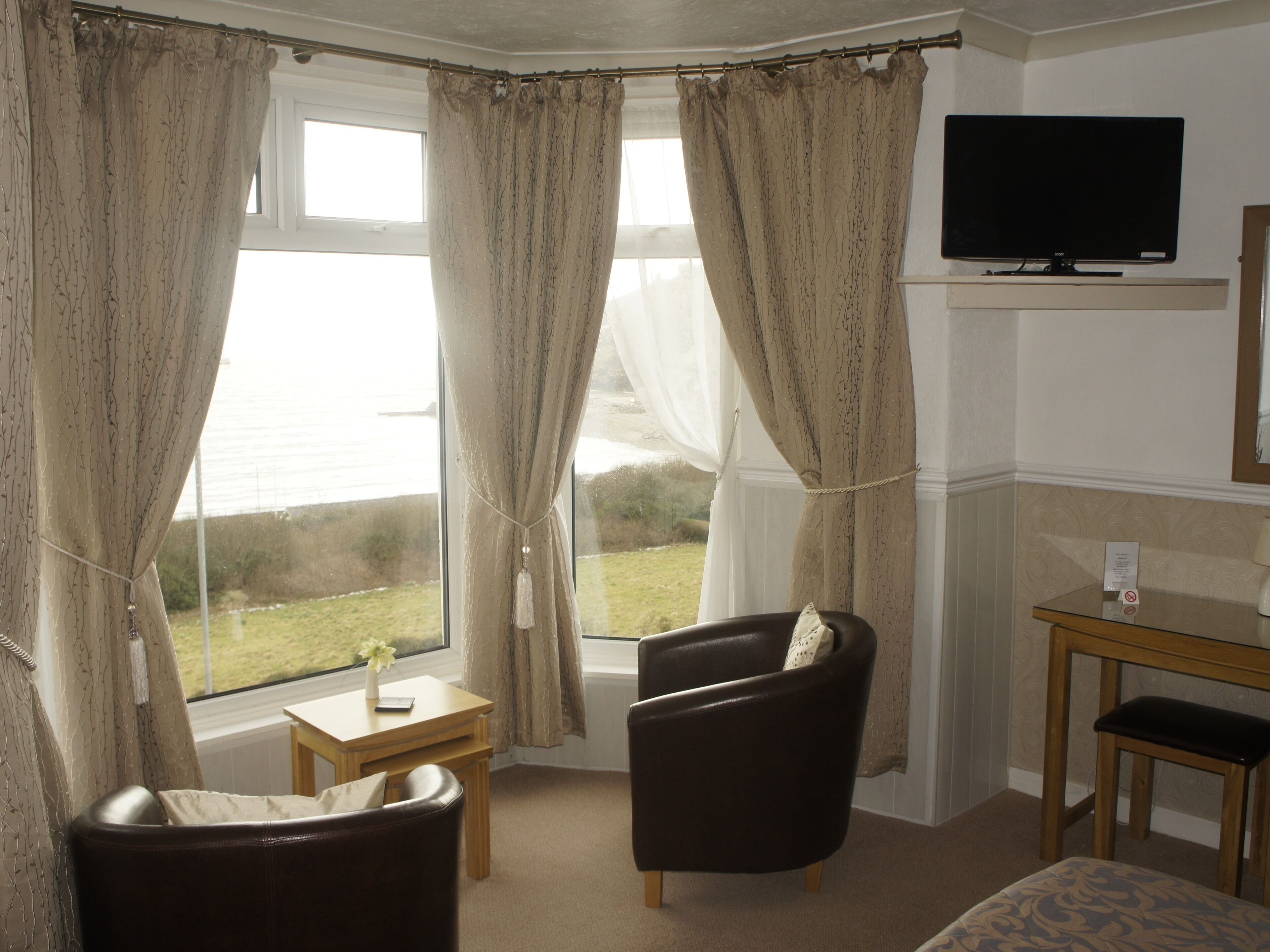 A room with a view of the sea, a TV on the wall, a desk, and two chairs.