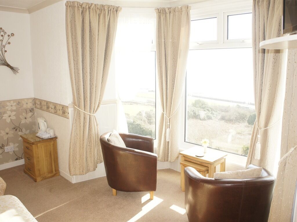 A room with two brown leather chairs, a small wooden table, a bedside table with a lamp, and a window with curtains.