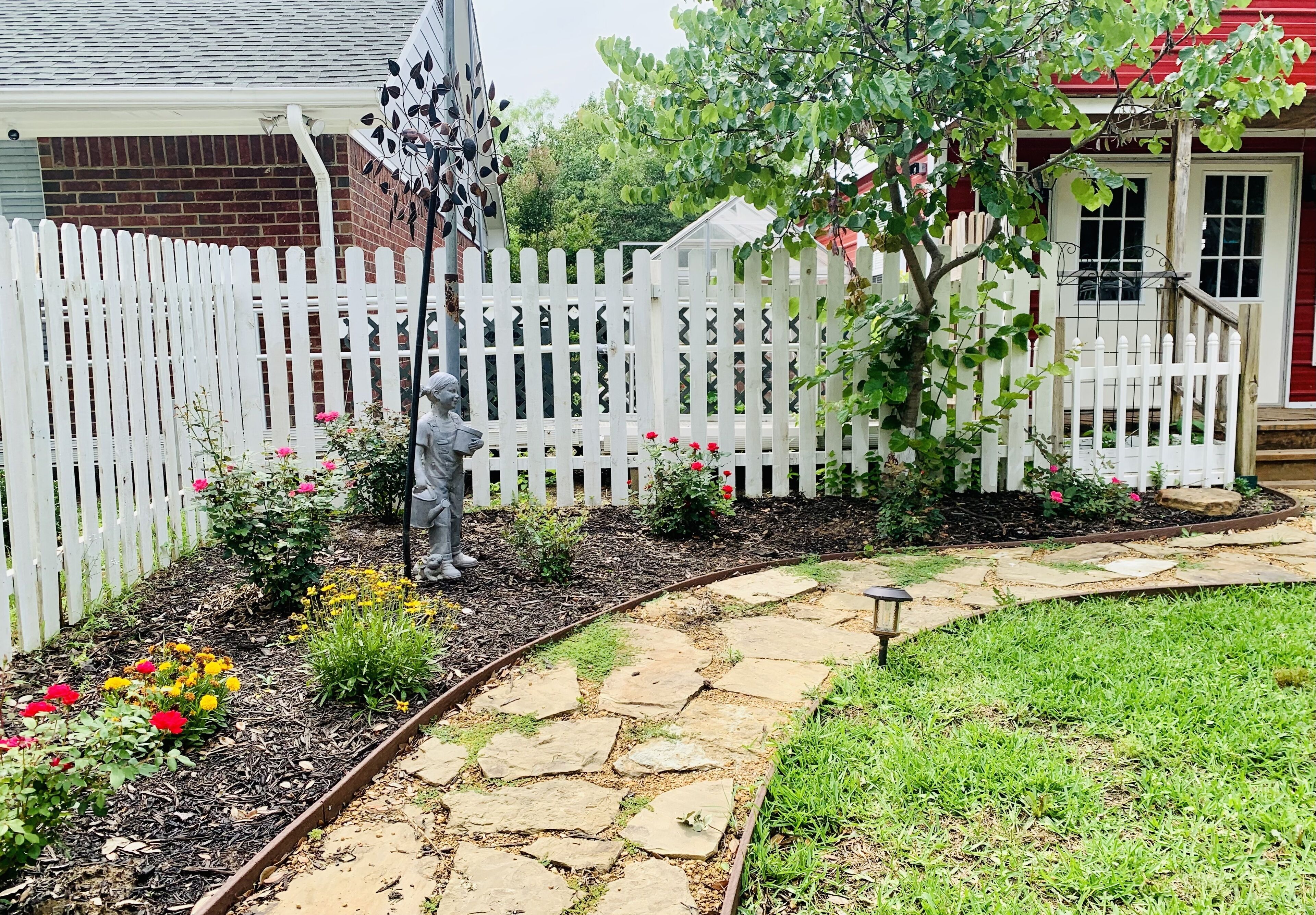 A garden with a white picket fence, a stone pathway, and various flowering plants.