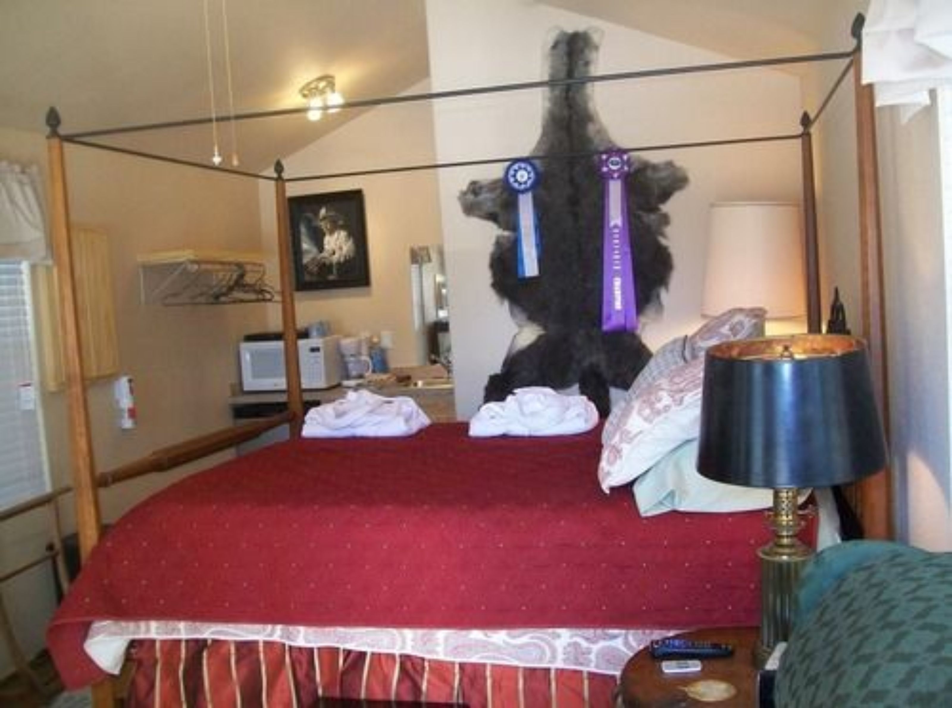 A four-poster bed with a red bedspread and white lace trim, a bedside lamp, and a small table with a lamp.