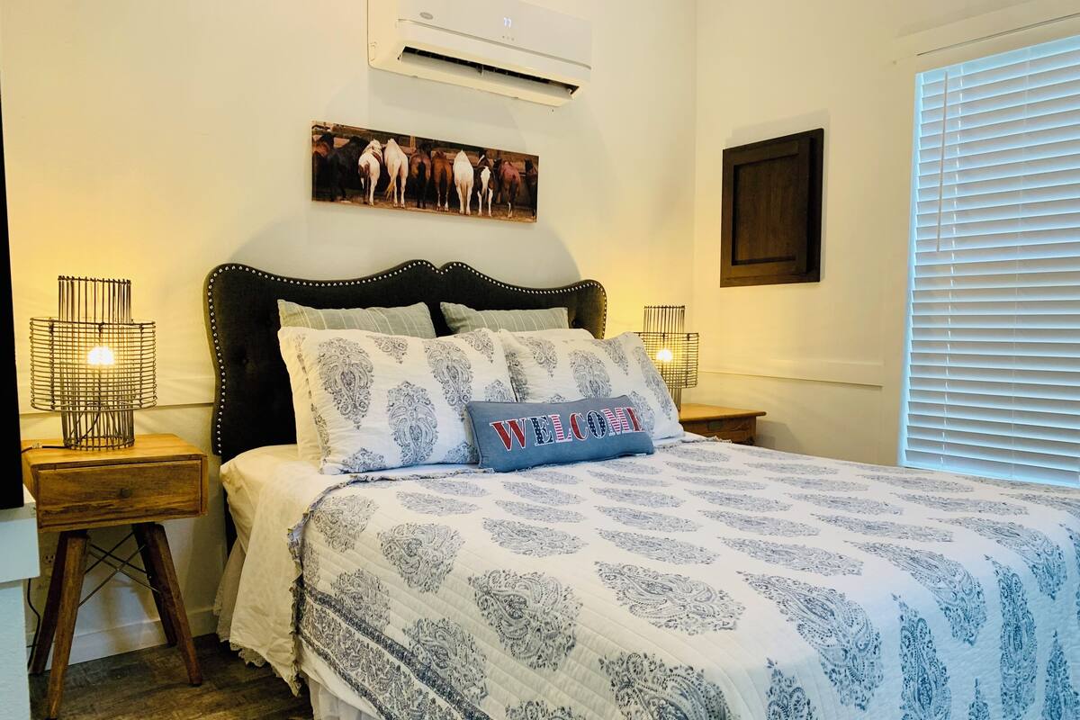 A bedroom with a bed, a headboard, a wall-mounted air conditioner, a picture, and a window with blinds.