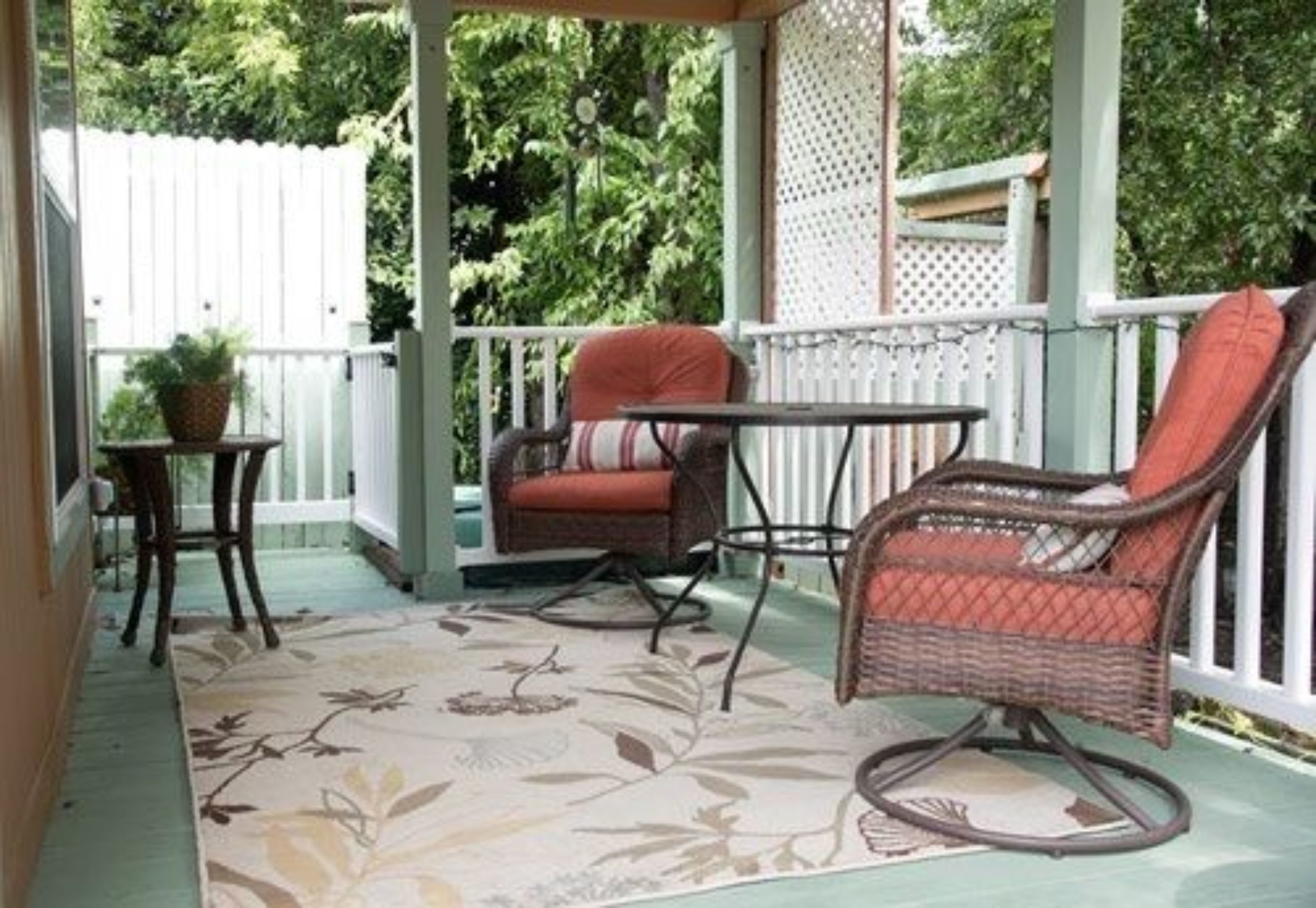 A covered porch with wicker chairs and a small table, surrounded by a white fence and lush greenery.