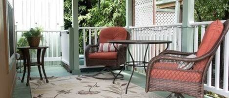 A covered porch with wicker chairs and a small table, surrounded by a white fence and lush greenery.