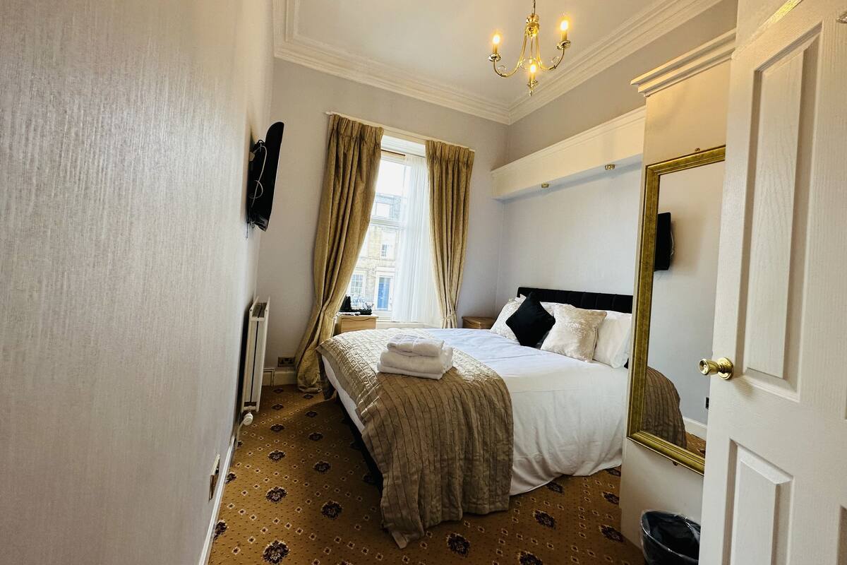 A hotel room with a large bed, a chandelier, a window with curtains, and a mirror on the door.