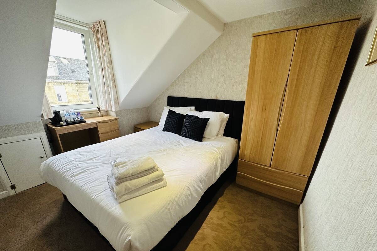 A neatly made bed with white linens and a folded towel, a wooden wardrobe, a bedside table with a lamp, and a window with curtains.