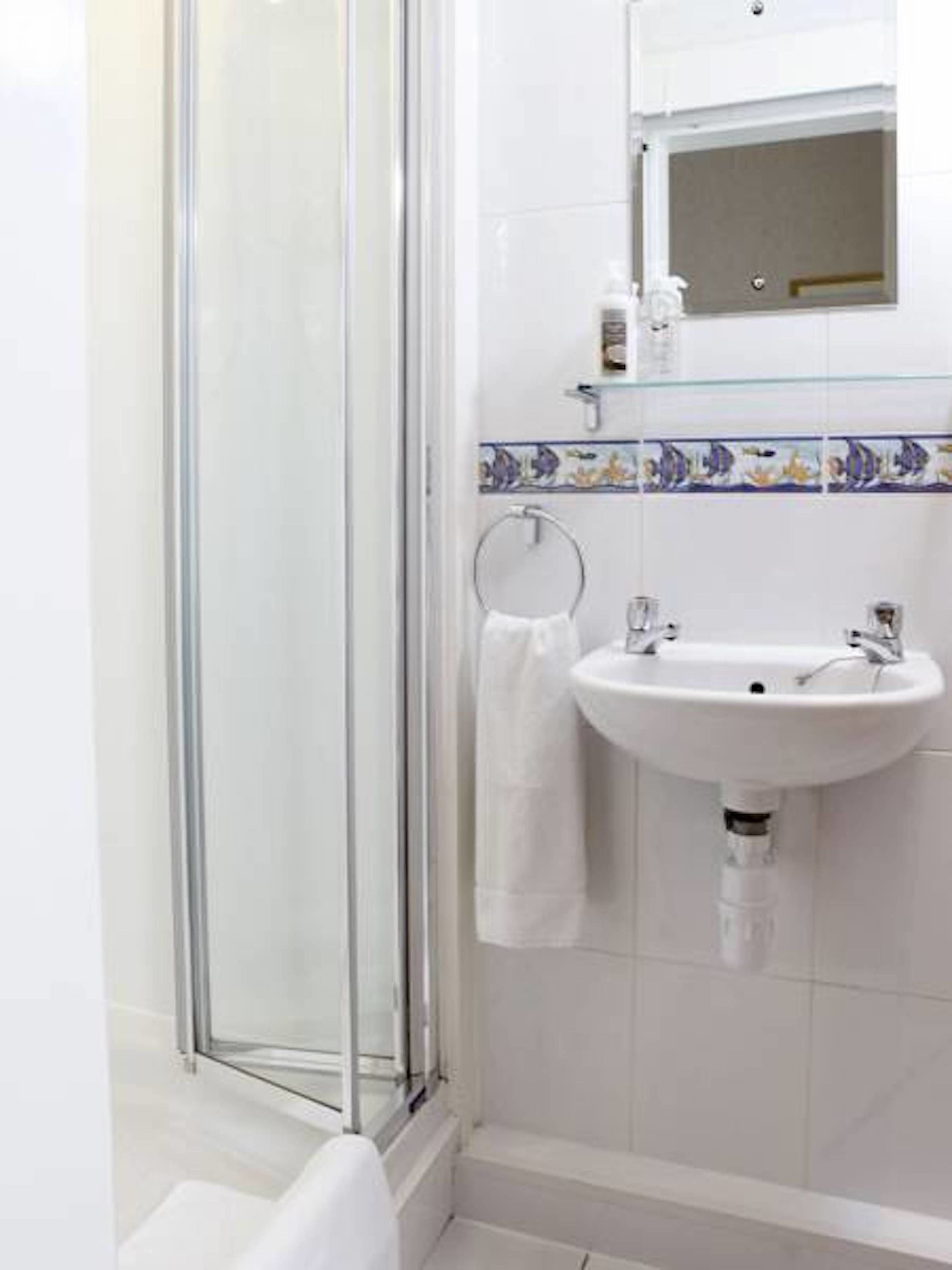 Une salle de bain dotée d’une douche à parois de verre, d’un lavabo blanc avec un miroir au-dessus, et d’un mur recouvert de carreaux avec une bordure décorative.
