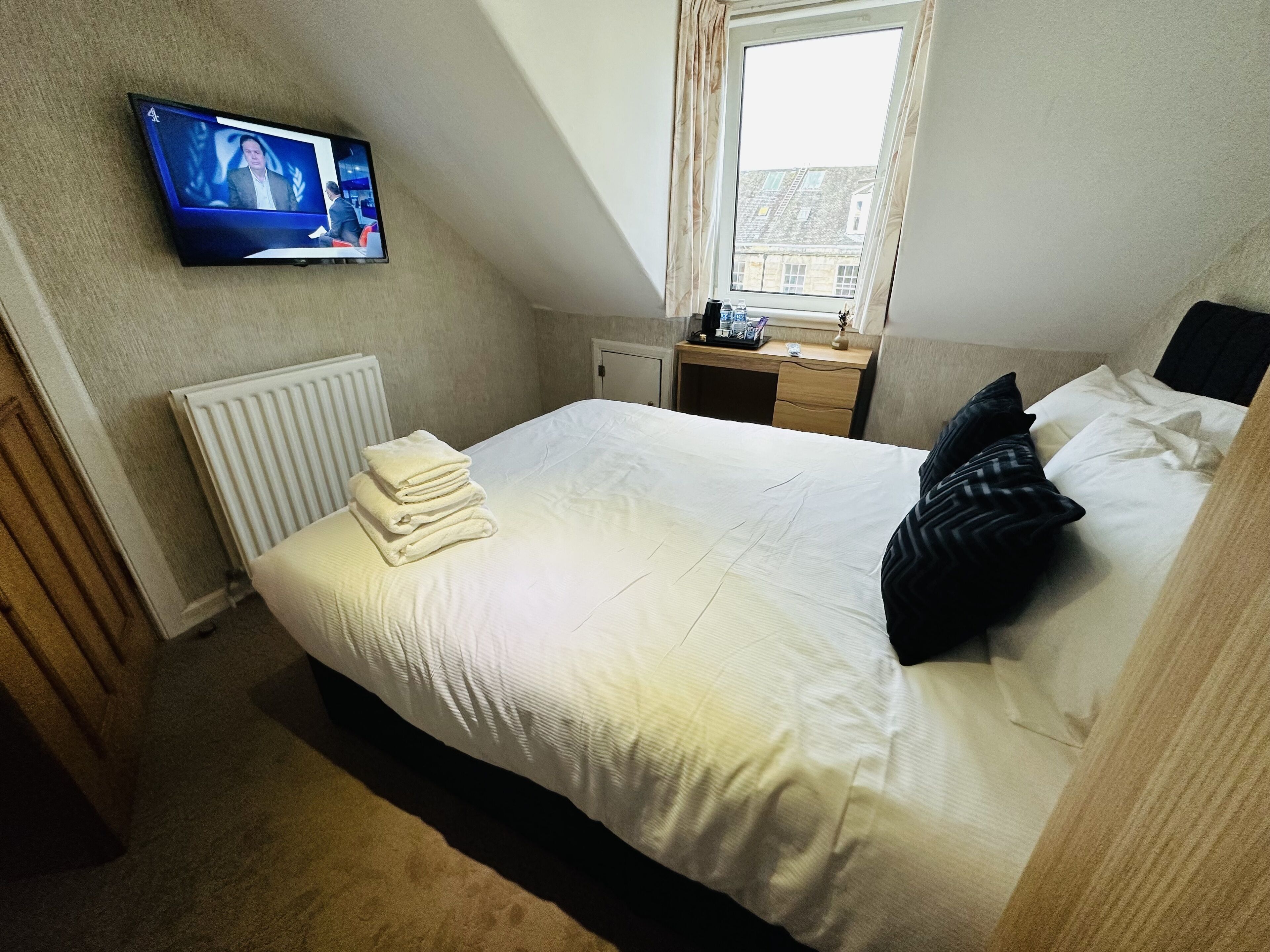 A hotel room with a bed, a TV mounted on the wall, a window with curtains, a desk with a lamp, and a radiator.