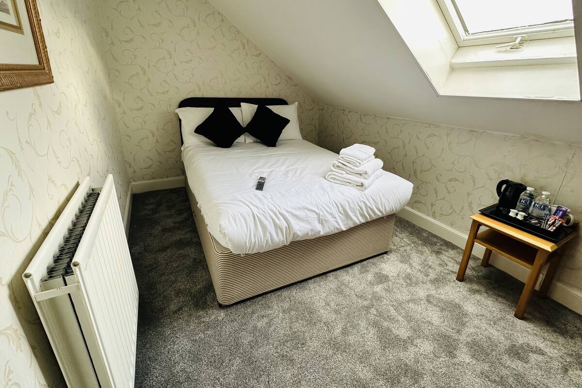 A small, neatly made bed with white linens and black pillows, a radiator, a small wooden side table with toiletries, and a window with a view of the sky.