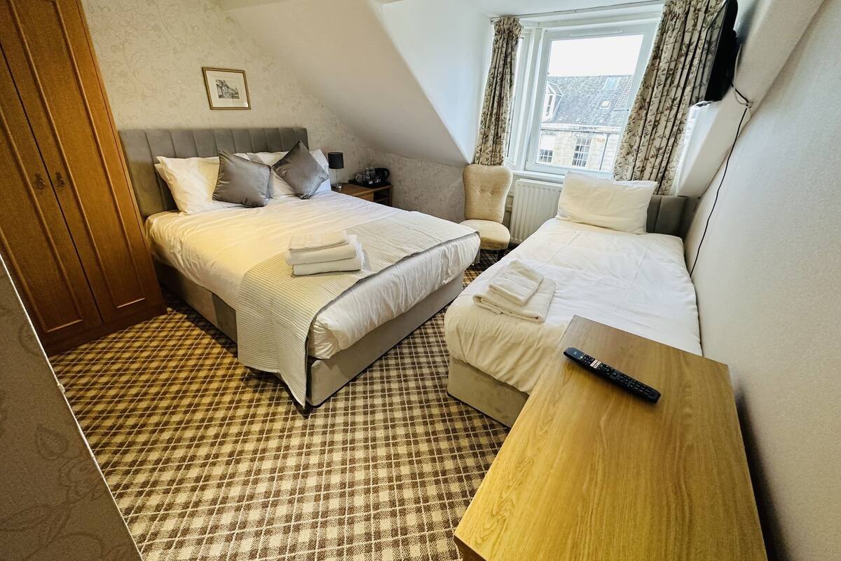 A hotel room with a large bed, a smaller bed, a wooden desk, a chair, a wardrobe, and a window with curtains.